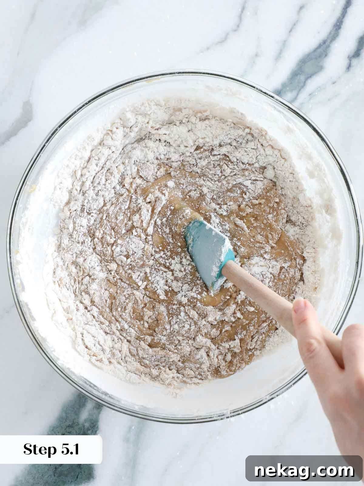A rubber spatula gently folding flour into the blondie batter in a mixing bowl, ensuring careful mixing.