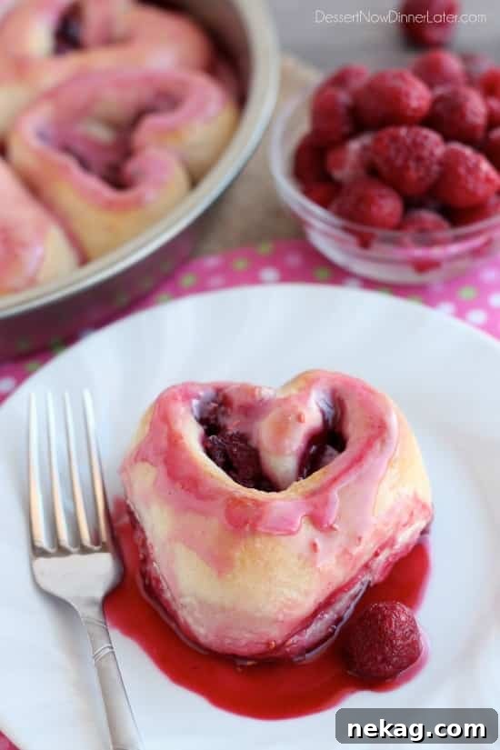 Sweetheart Raspberry Swirls 3 A single heart-shaped raspberry roll on a white plate, showcasing its soft texture and the vibrant pink raspberry glaze drizzled generously over the top.