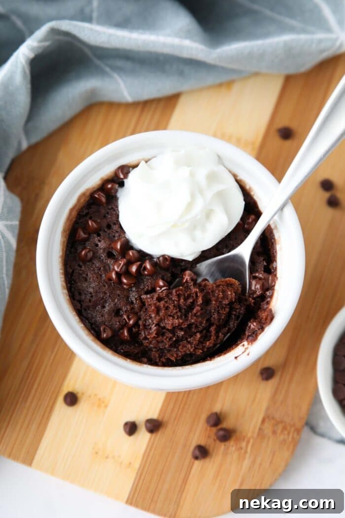 A perfectly portioned microwave chocolate cake, served in a small ramekin, generously topped with mini chocolate chips and a dollop of fresh whipped cream. A spoon has taken out a delicious bite, showing the cake's moist, fudgy texture.