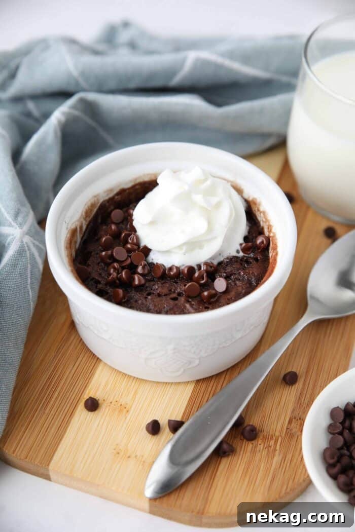 A close-up view of a rich microwave chocolate cake nestled in a ceramic ramekin, adorned with a generous sprinkle of mini chocolate chips and a swirl of light, airy whipped cream. The focus highlights the inviting texture and delicious toppings.