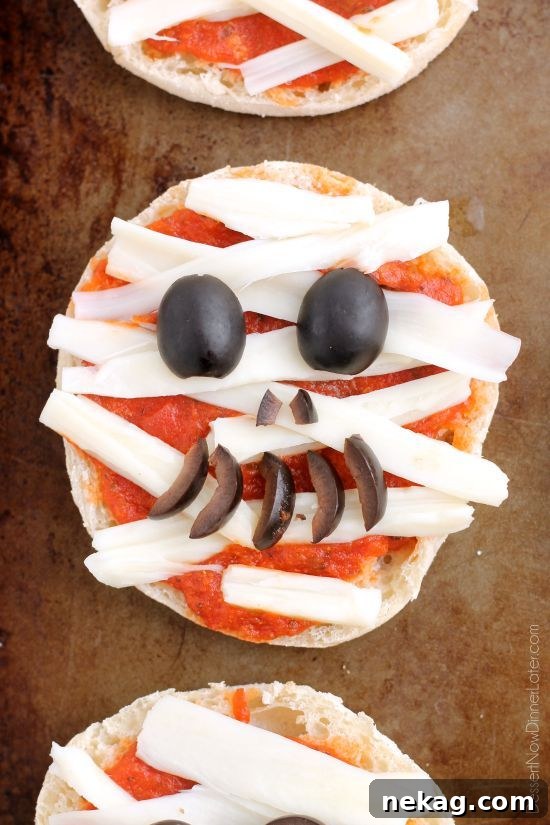 Spooky Swaddle Pizzettes 6 Another image of hands arranging black olive pieces to create a mummy's eyes.