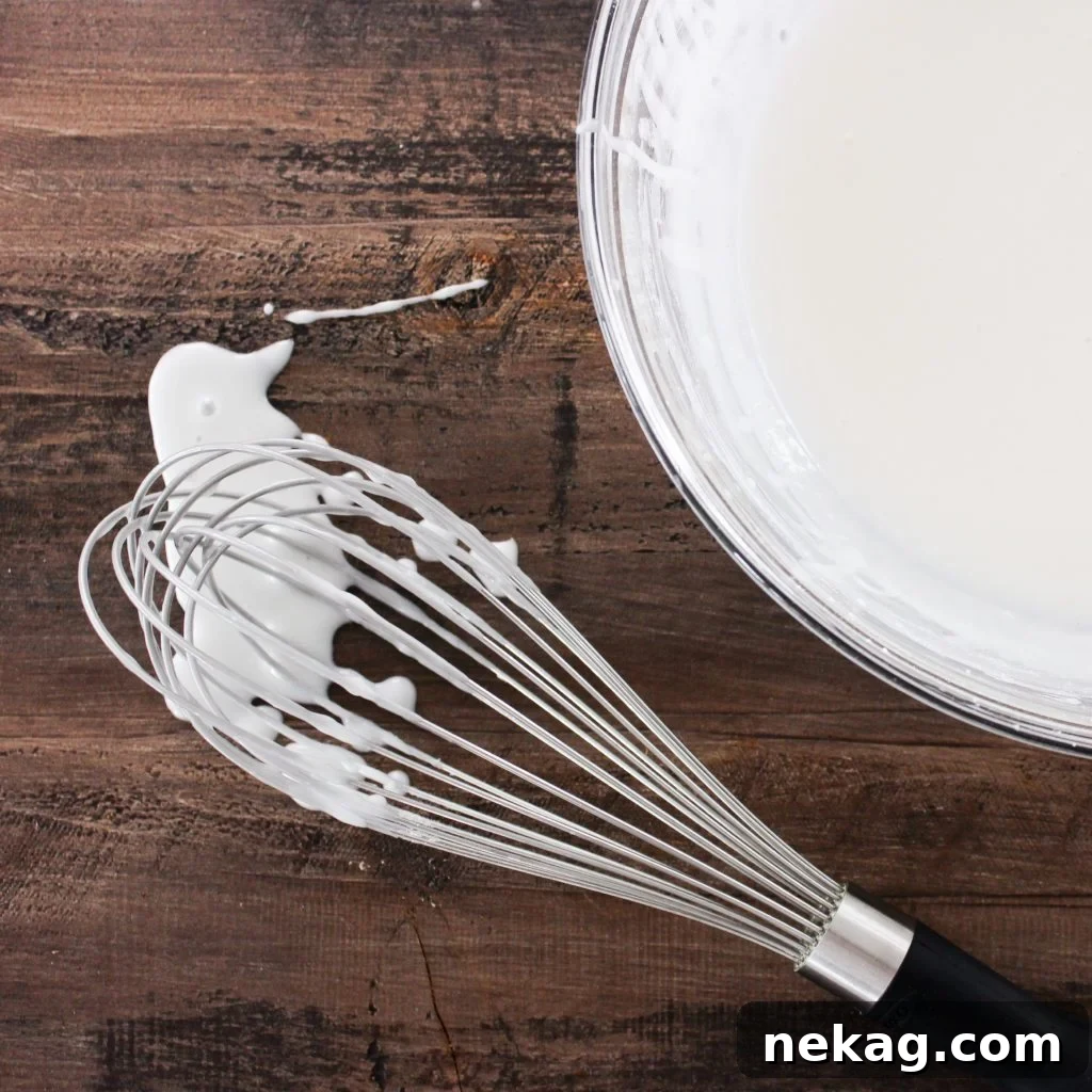 Irresistibly Delicious Donut Glaze 4 White glaze spilling from a whisk onto a wood board next to a bowl of white glaze, showcasing its ideal, pourable consistency for dipping donuts.
