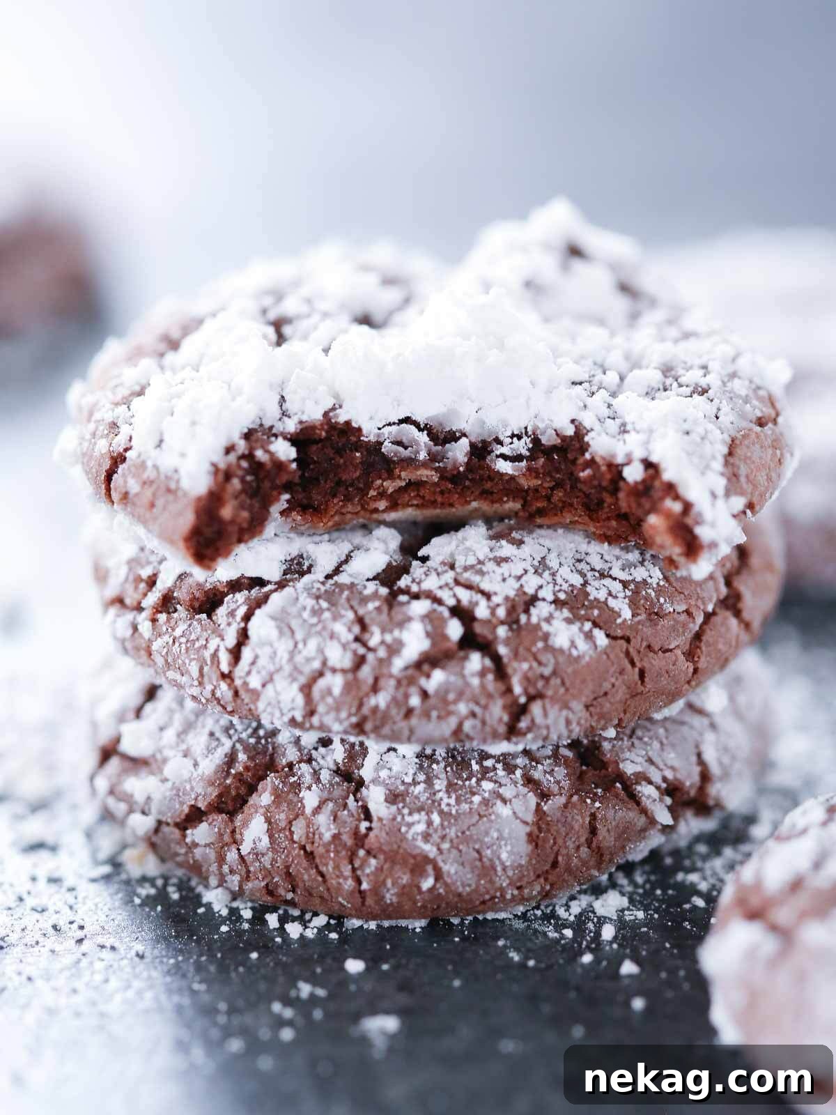 A stack of rich chocolate crinkle cookies, with the top one bitten to reveal its fudgy interior, covered in white powdered sugar.