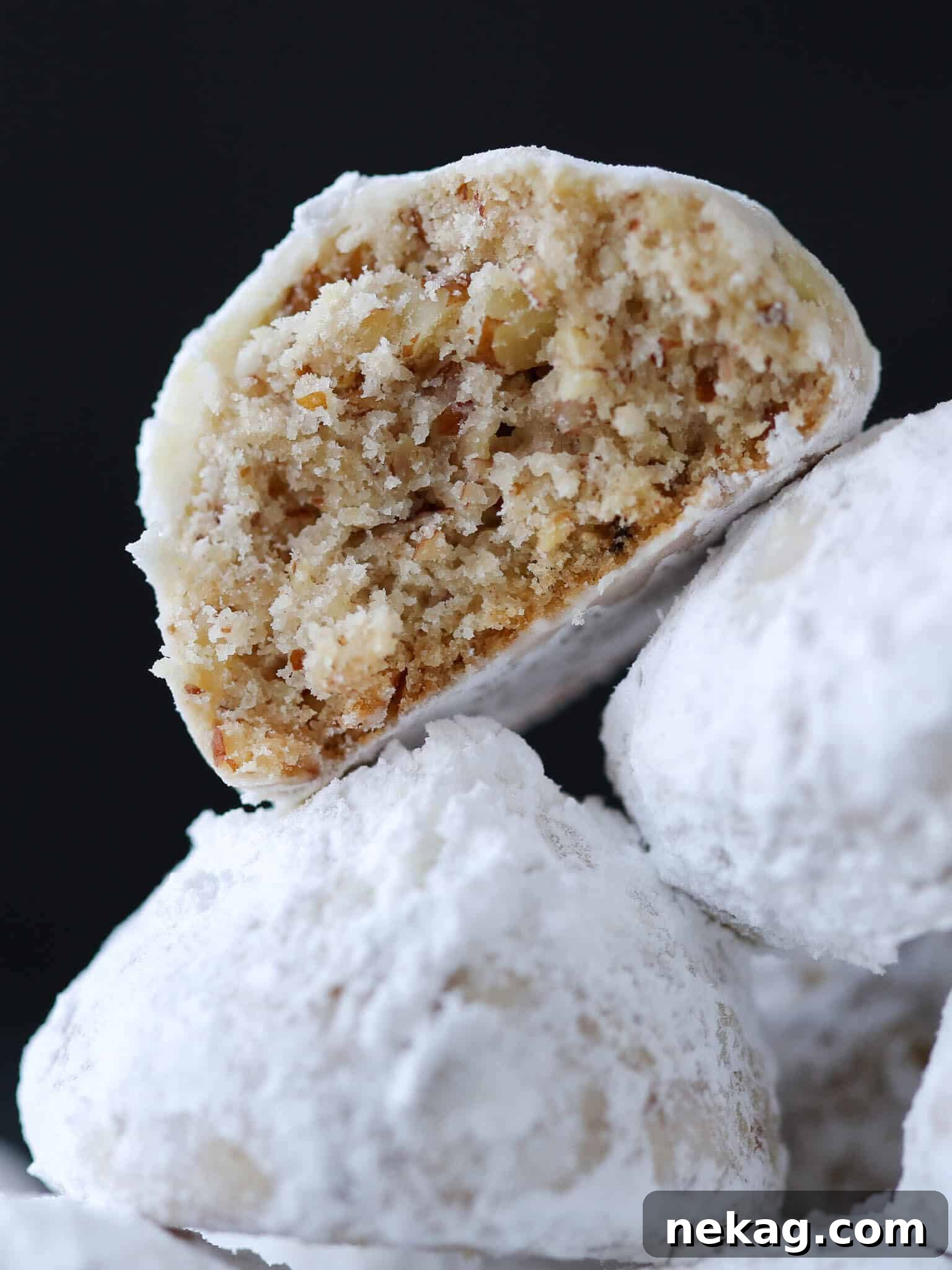 Buttery Pecan Snowball Bites 2 Three perfectly formed snowball cookies dusted with powdered sugar, with the top cookie broken open to reveal its crumbly, pecan-studded interior.