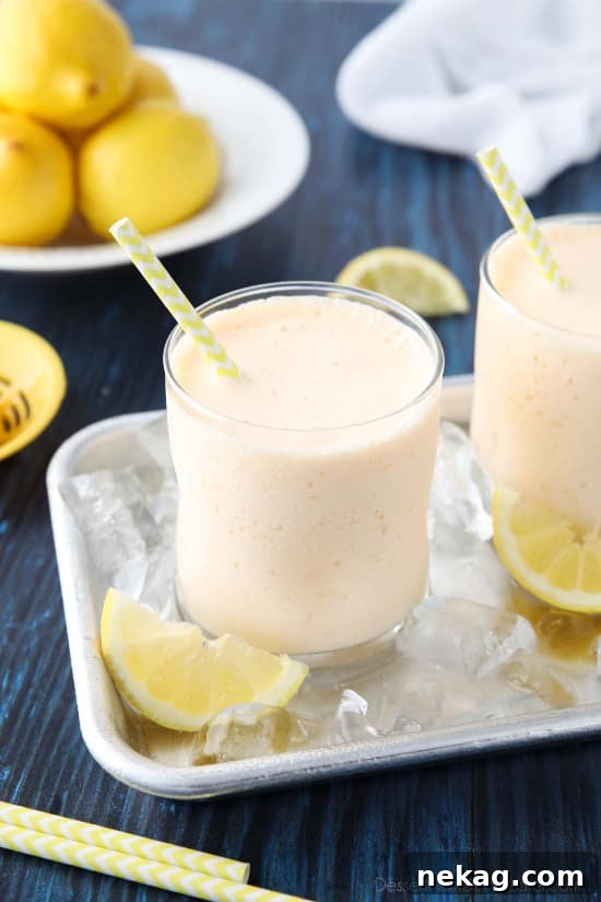 A tall glass of creamy, pale yellow frosted lemonade, garnished with a fresh lemon slice, sits on a wooden table, promising a refreshing summer treat.