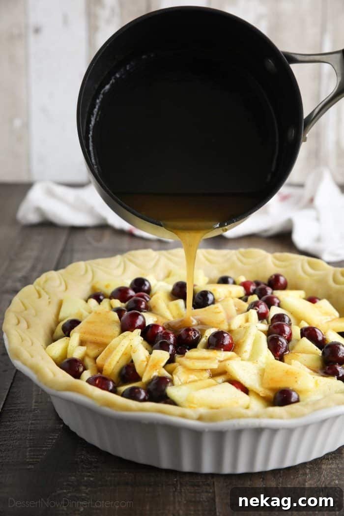 Syrup being generously poured over the cranberry and sliced apple filling in an unbaked pie crust, highlighting the glossy texture.
