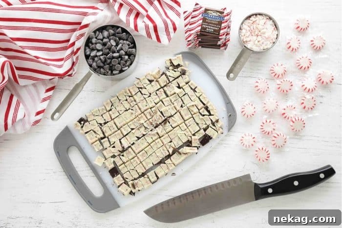 A wooden cutting board with expertly chopped peppermint bark squares and a chef's knife, flanked by measuring cups brimming with semi-sweet chocolate chips and perfectly crushed peppermint candies, highlighting fresh ingredients.