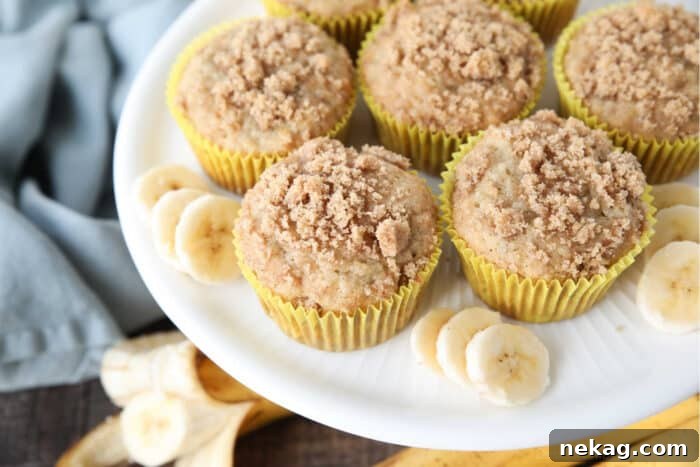 Golden Banana Streusel Muffins 9 White cake plate with banana muffins in yellow wrappers with crumb streusel on top and slices of banana on the sides.