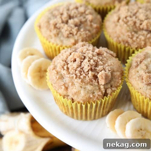 White cake plate with banana muffins in yellow wrappers with crumb streusel on top and slices of banana on the sides.