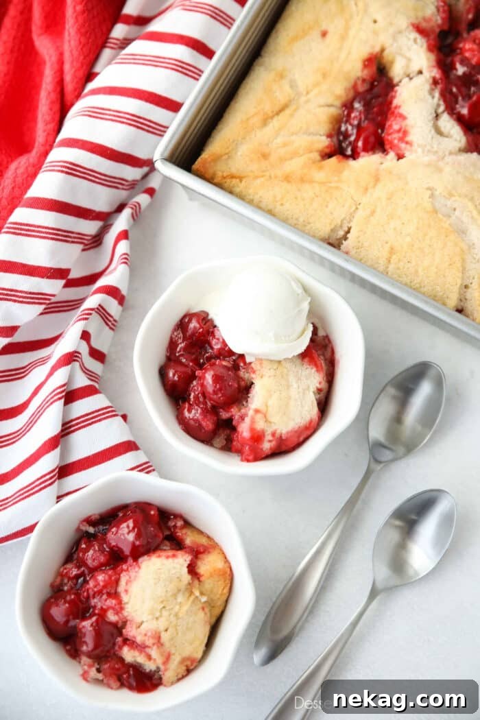 Two bowls of easy cherry cobbler with two spoons on the side. One serving of cobbler has ice cream on top, and is sitting next to the pan it was baked in.