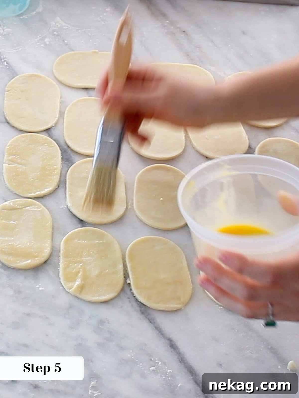 Pie dough shapes arranged on a baking sheet, with egg wash being applied to the edges of one half.