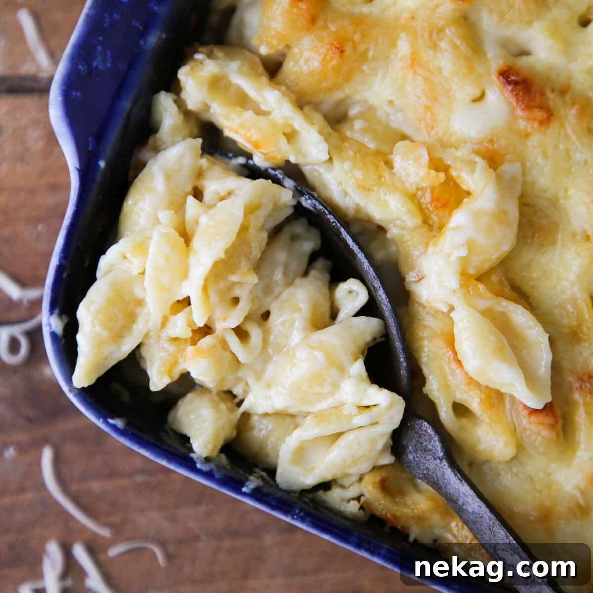 A serving spoon lifting a generous portion of hot, creamy white cheddar mac and cheese from a rustic blue baking dish on a wooden surface.