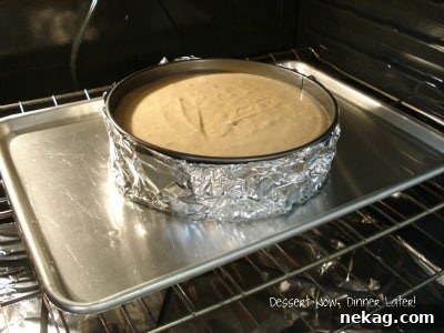 Perfect Gingersnap Pumpkin Cheesecake Minus The Cracks 6 A springform pan with cheesecake baking in a water bath, placed inside a larger rimmed baking sheet in the oven.