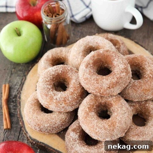 Baked Apple Cider Donuts stacked on top of a wooden cutting board with a cup of cider, fresh apples, and cinnamon sticks on the side.