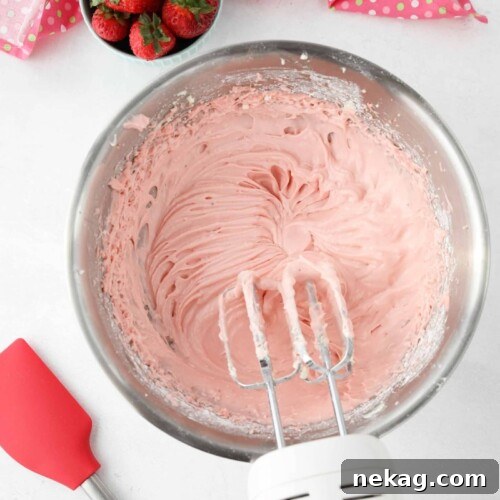 A close-up of a bowl filled with smooth, pink strawberry cream cheese frosting, with an electric mixer and beaters nearby, ready to frost your favorite treats.