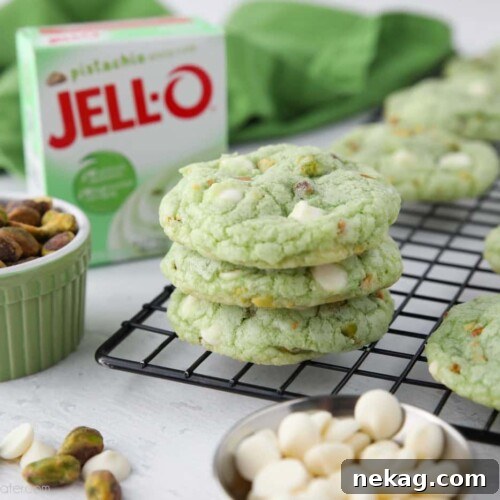 Stack of pistachio pudding cookies on a cooling rack with a jello pudding box, pistachio nuts, and white chocolate chips nearby.