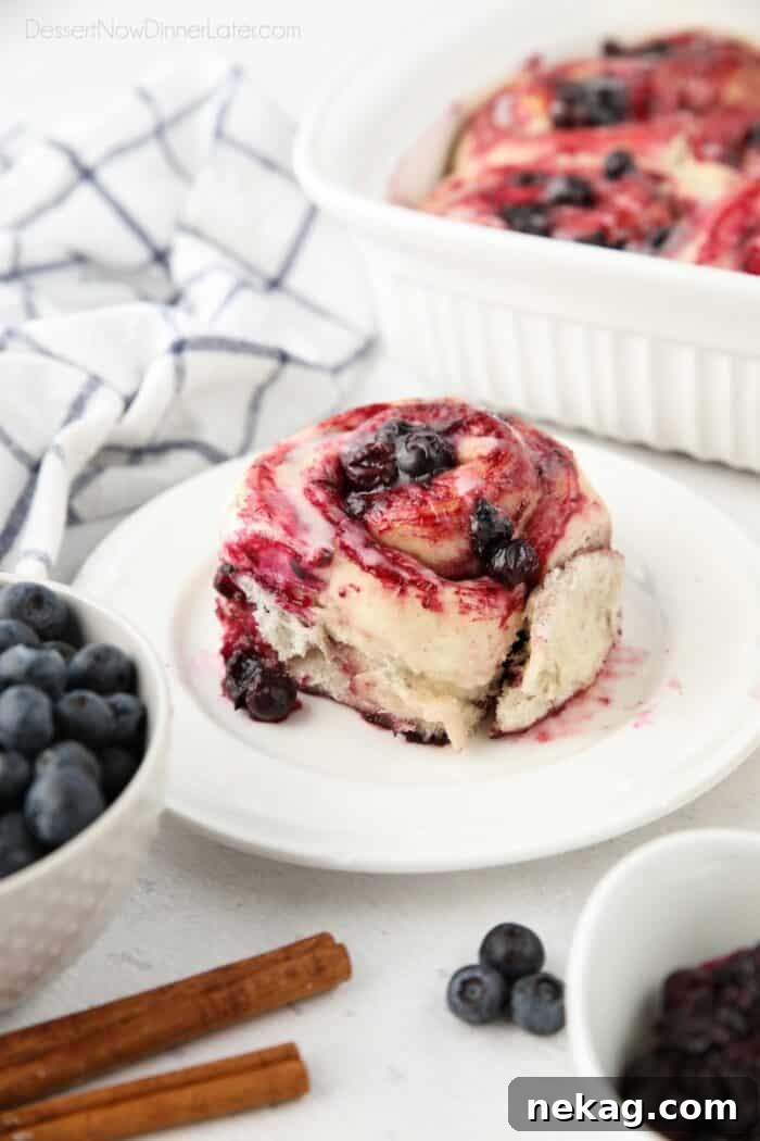 Gooey Blueberry Cinnamon Swirls 2 A single, perfectly golden-brown blueberry cinnamon roll on a white plate, adorned with glistening lemon glaze and fresh blueberries, highlighting its soft texture and rich filling.