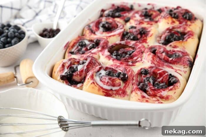 Gooey Blueberry Cinnamon Swirls 14 An angled view of a casserole dish brimming with freshly baked blueberry cinnamon rolls, generously coated in a shimmering lemon glaze and sweet blueberry sauce, ready to be served.