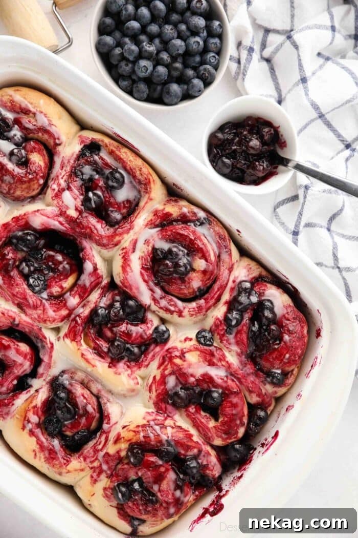 Gooey Blueberry Cinnamon Swirls 3 A baking dish filled with twelve freshly baked blueberry cinnamon rolls, each topped with a delicate lemon glaze and a scattering of fresh blueberries, viewed from above.