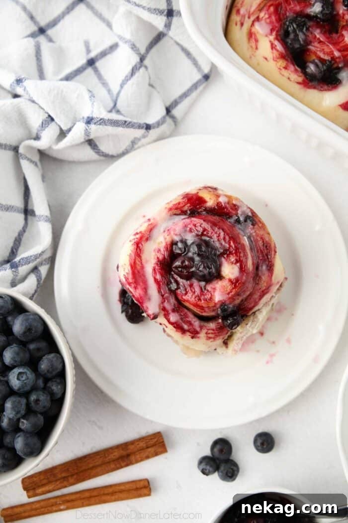 Gooey Blueberry Cinnamon Swirls 4 Close-up, top-down view of a single blueberry cinnamon roll on a clean white plate, showcasing the spiraled layers of dough, blueberry sauce, and smooth lemon glaze.