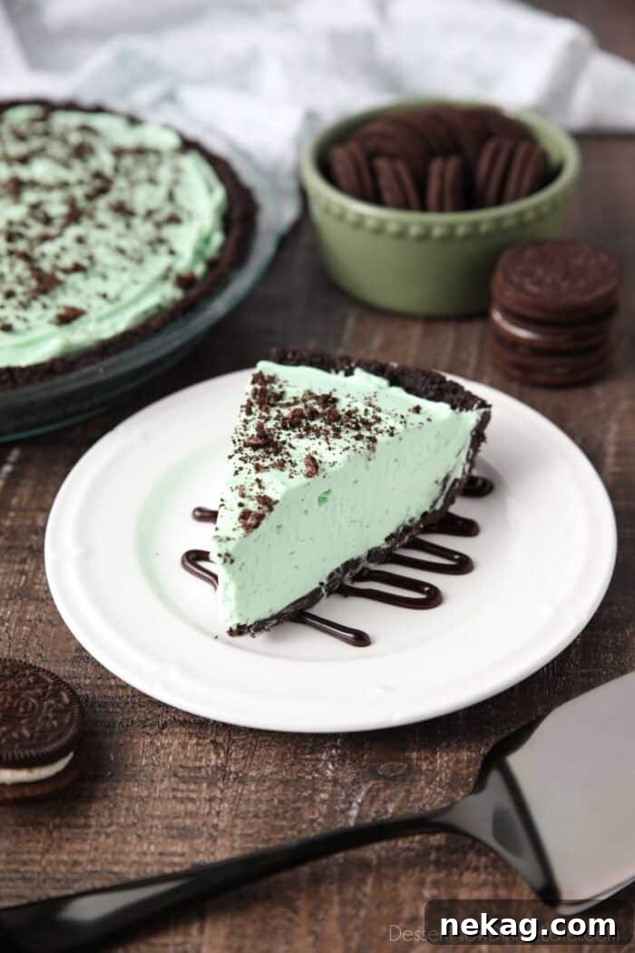 A perfectly sliced piece of mint grasshopper pie with a dark Oreo crust, drizzled with chocolate sauce on a white plate.
