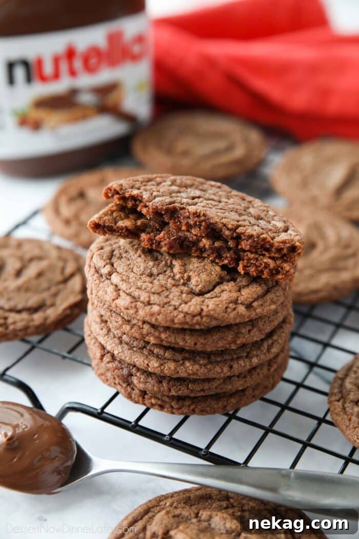 A Nutella cookie broken in half, revealing its soft, fudgy interior with chocolate hazelnut goodness, stacked on top of more freshly baked Nutella cookies.