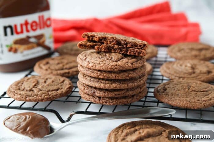 A Nutella cookie broken in half, showcasing its perfect fudgy center, with more cookies artfully arranged in the background.