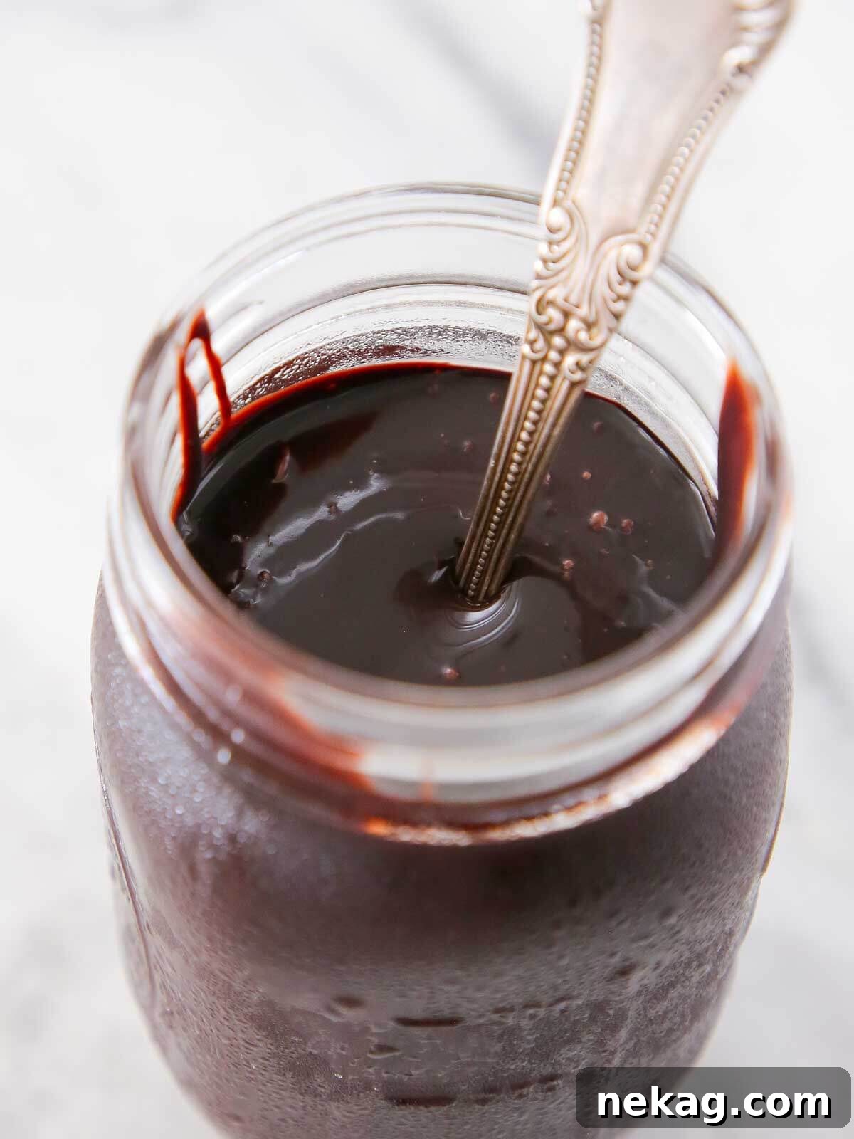 A glass jar filled with luscious, dark chocolate hot fudge sauce, with a spoon resting in it.