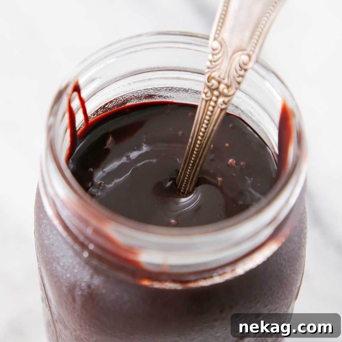 Three glass jars of chocolate fudge sauce, one with a spoon, arranged on a marble surface.