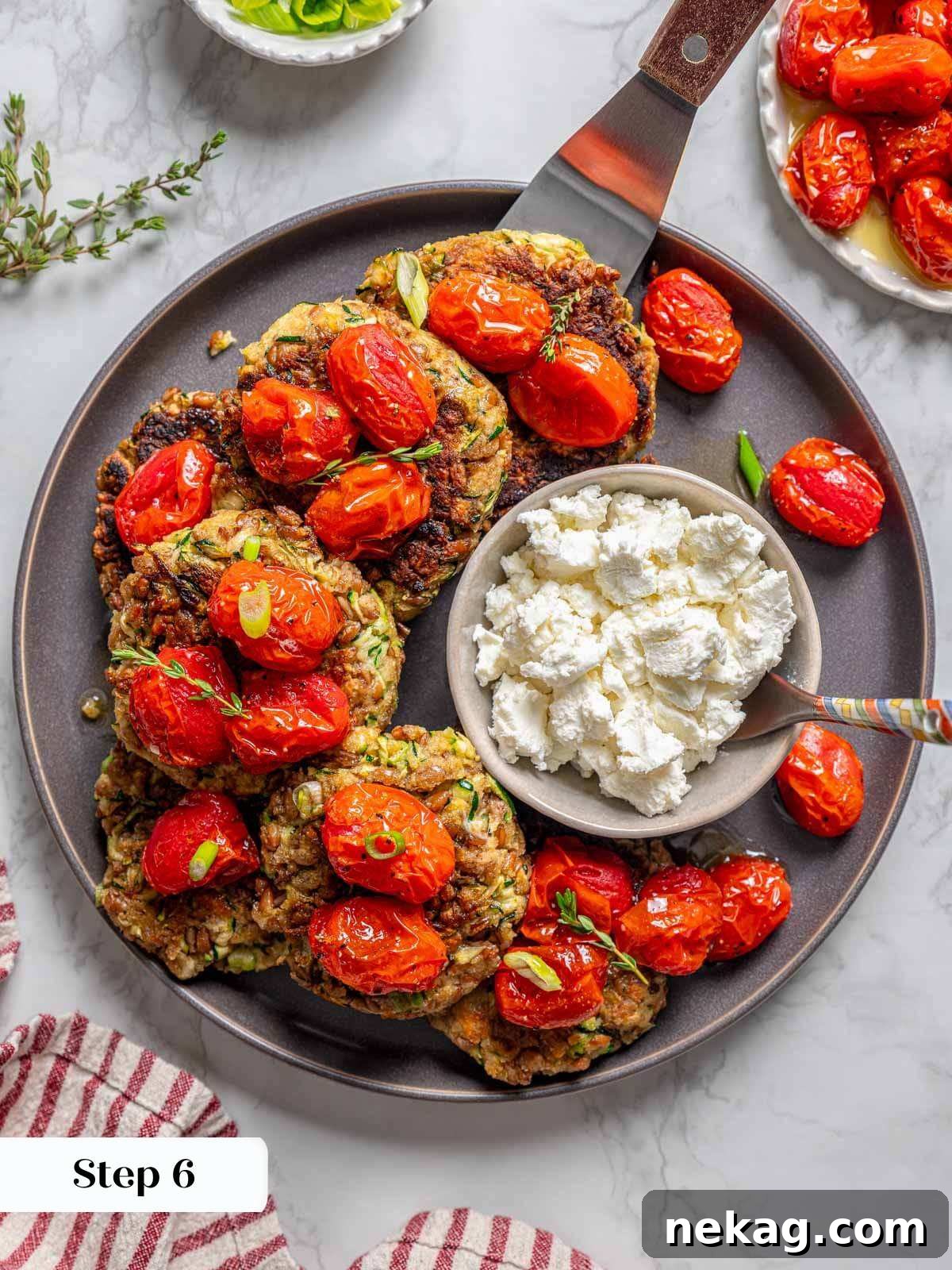 A serving of beautifully assembled zucchini fritters, topped with tomatoes and goat cheese, on a grey plate with a spatula for serving.