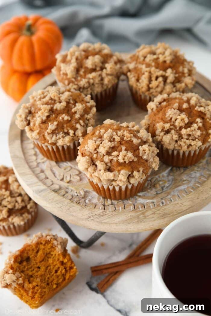 A trio of pumpkin streusel muffins, perfectly baked with abundant crumb topping, resting on a white cake stand against a warm background.