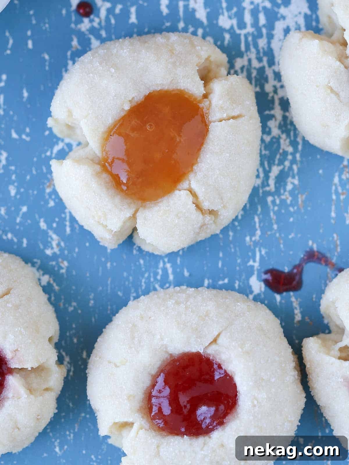 Preserve-Kissed Shortbreads 11 Two thumbprint cookies, one with strawberry and one with apricot jam, on a blue surface.