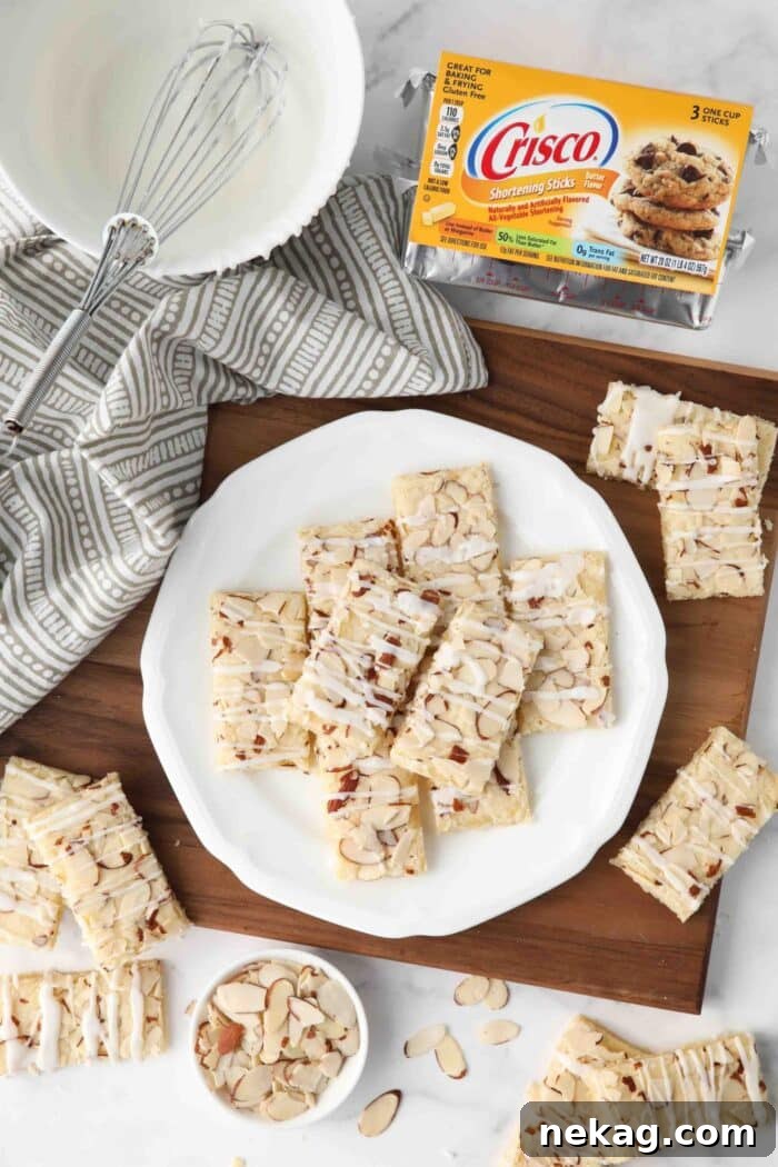 Top view of almond bars on a plate with Crisco shortening, sliced almonds, and a bowl of glaze on the side.