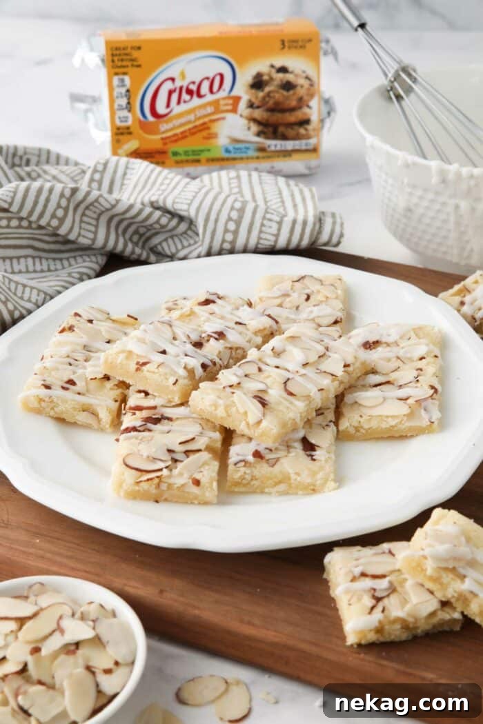 Side view of almond bars on a plate with Crisco shortening, sliced almonds, and a bowl of glaze nearby.