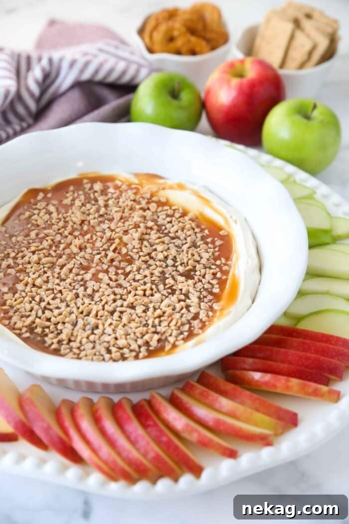 A beautifully presented bowl of creamy caramel apple dip, artfully garnished with more caramel drizzles and toffee, with a fan of sliced red and green apples beside it.