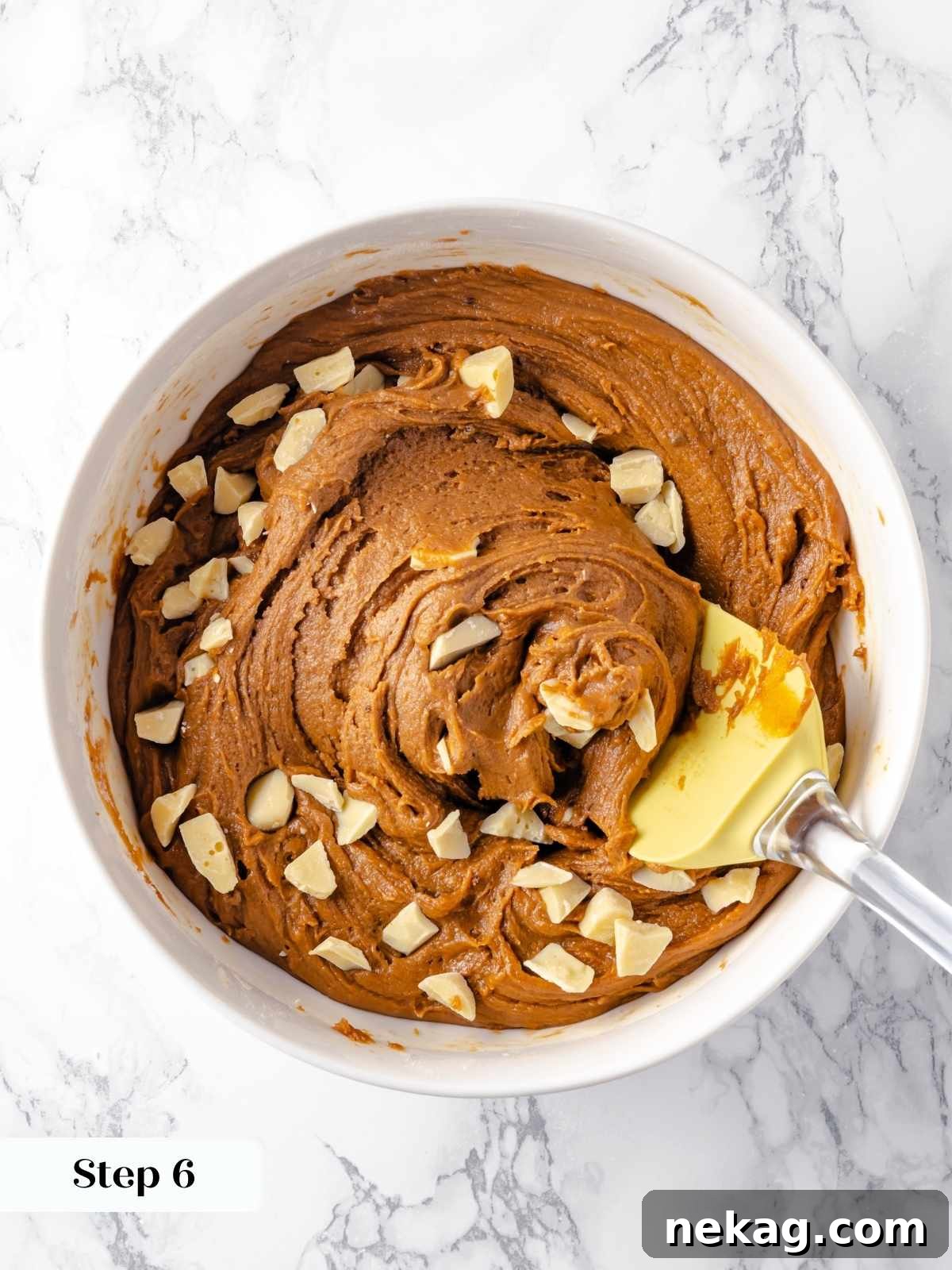 Festive Gingerbread Blondies 10 A mixing spatula expertly folds creamy white chocolate chips into the spiced gingerbread blondie batter within a clear glass bowl, ensuring even distribution.
