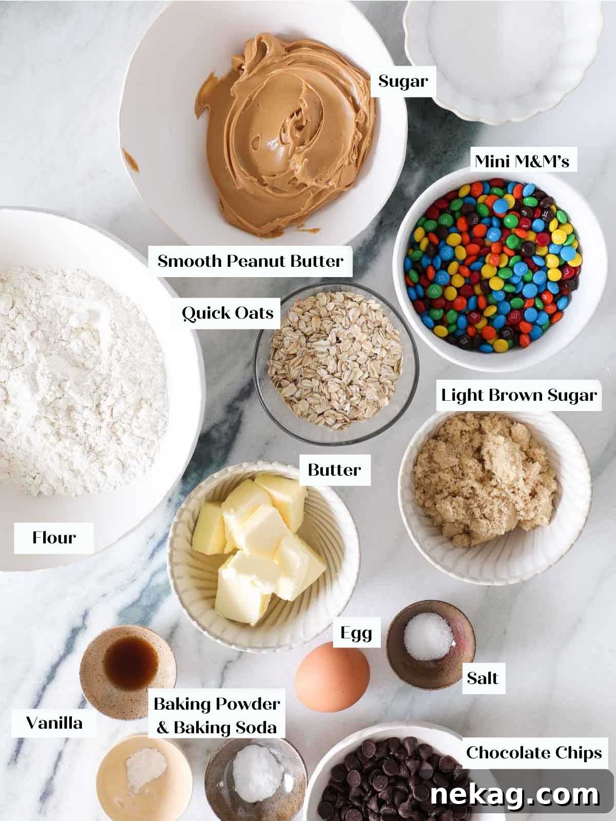 A flat lay photograph of various monster cookie ingredients neatly arranged on a white marble counter, featuring oats, creamy peanut butter, and colorful M&Ms, ready for baking.