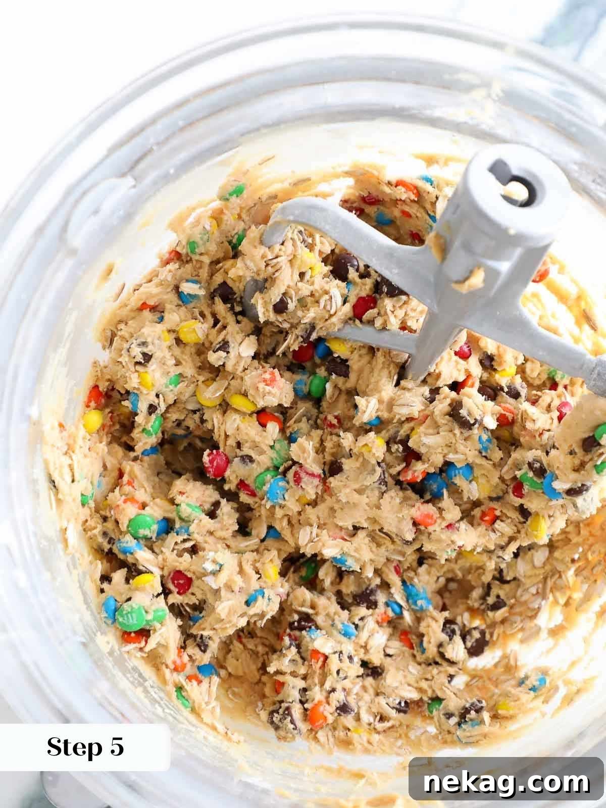 A clear mixing bowl brimming with chunky monster cookie dough, generously packed with chocolate chips, M&Ms, and oats.