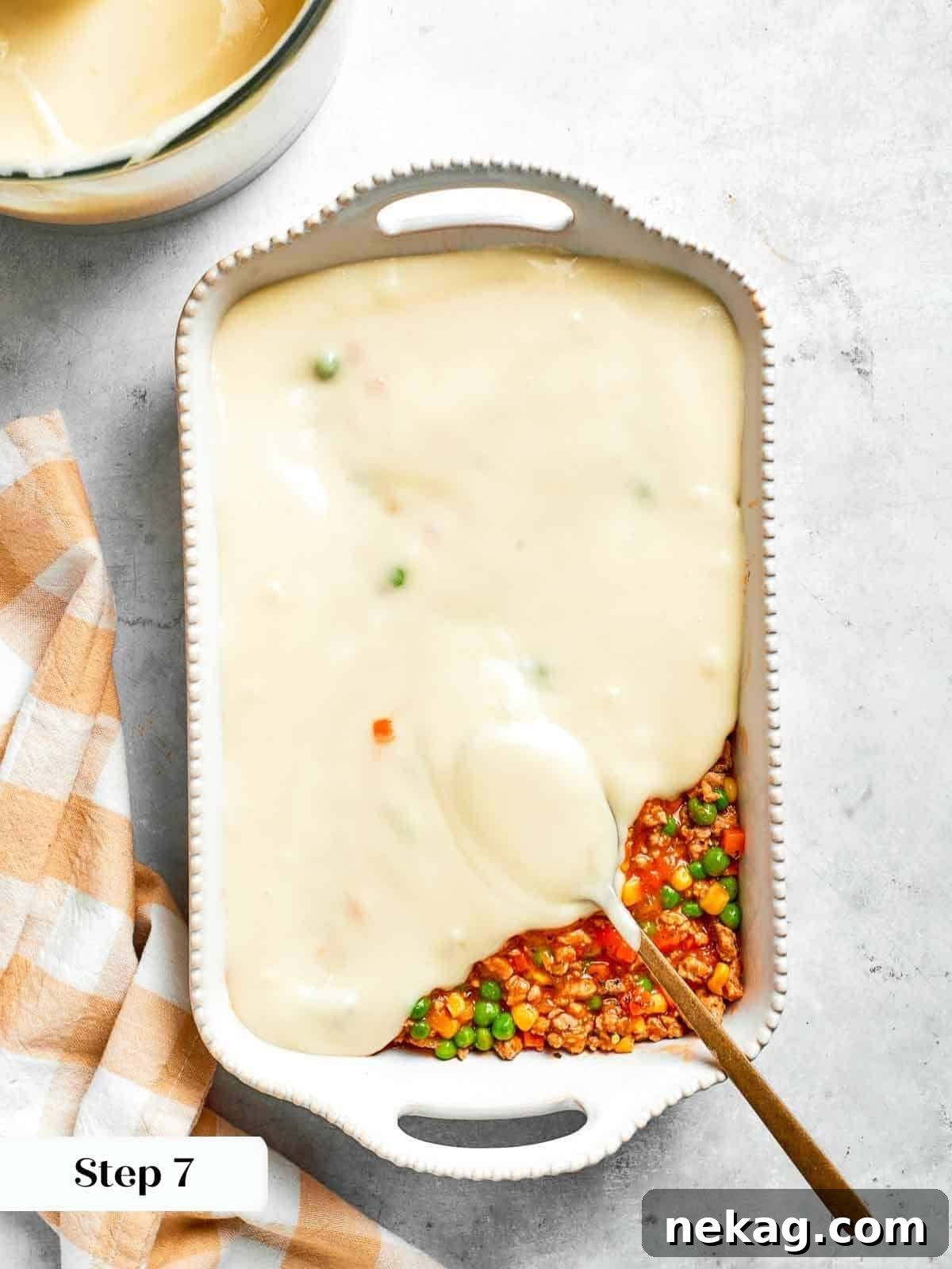 A spoon gently and evenly layering mashed potatoes over the rich chicken and vegetable filling, preparing the pie for its final bake.