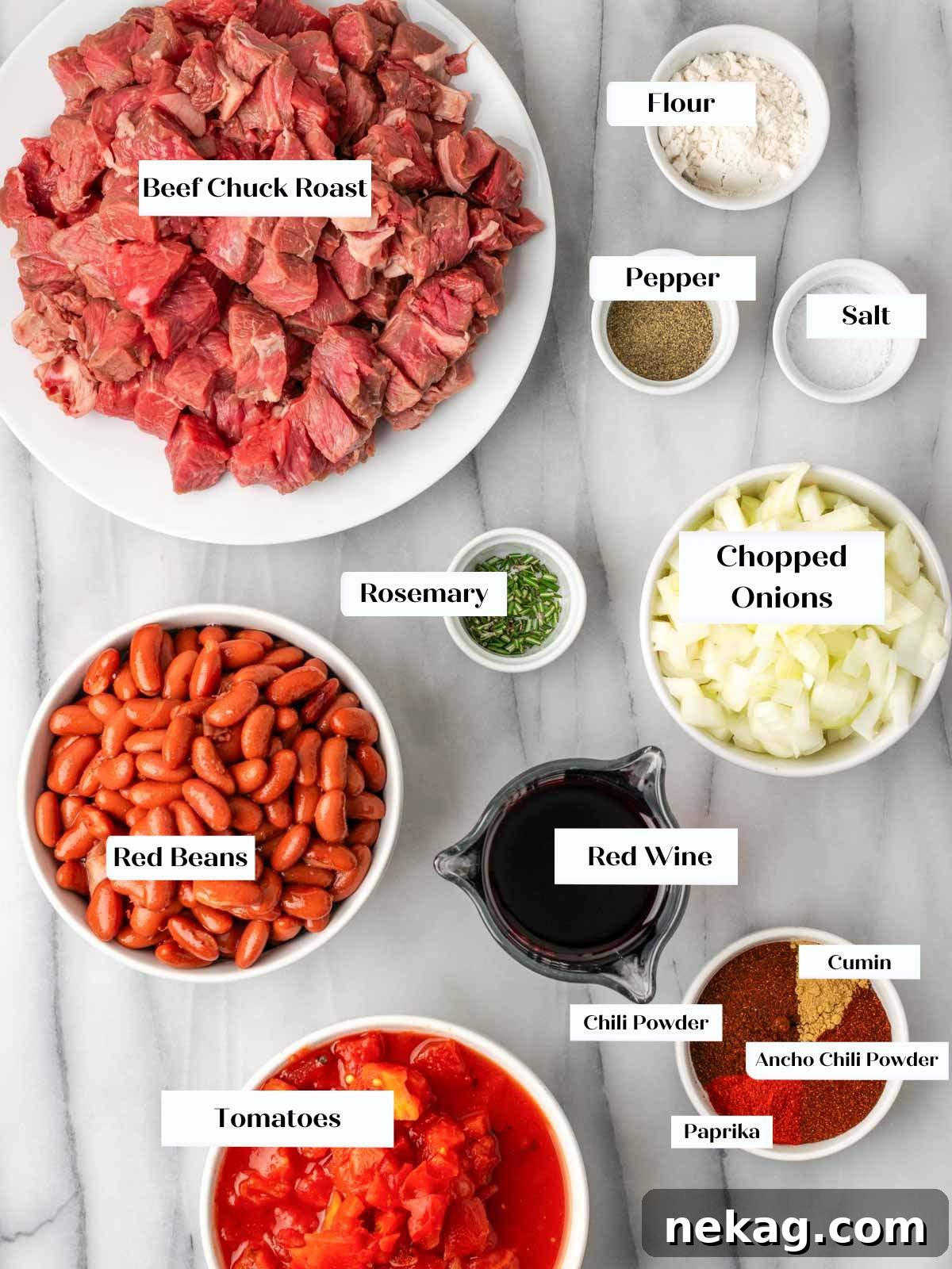 Assorted steak chili ingredients neatly arranged on a marble countertop, including beef chunks, onions, various spices, cans of beans and tomatoes, and a sprig of fresh rosemary.