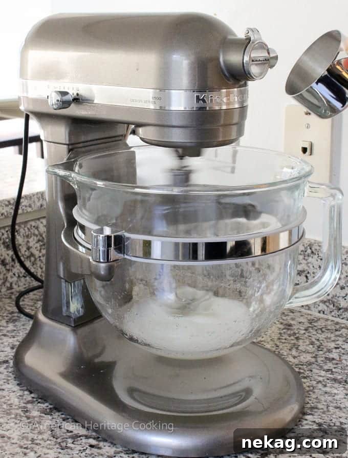 A silver stand mixer with a clear bowl and egg whites at the bottom, ready for whipping.
