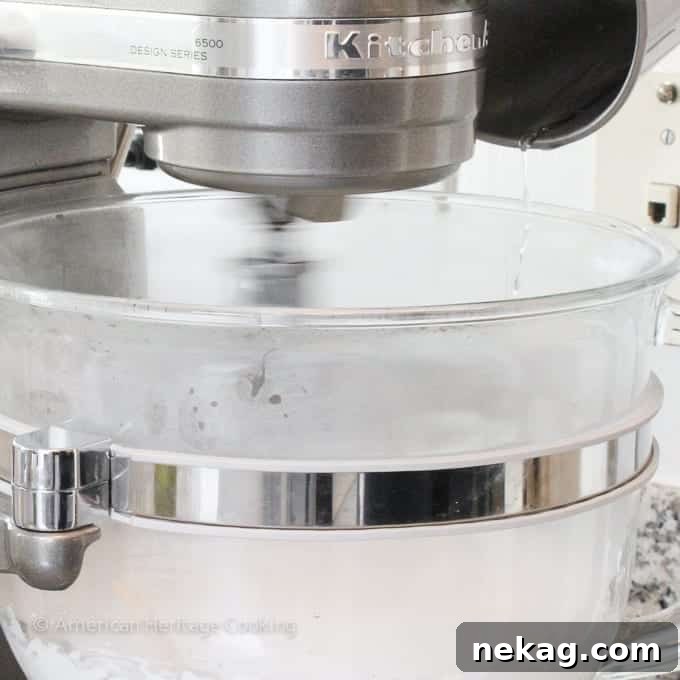 The whisk attachment of a stand mixer blurred to show it's in motion, with a thin stream of sugar syrup being poured down the side of the bowl.