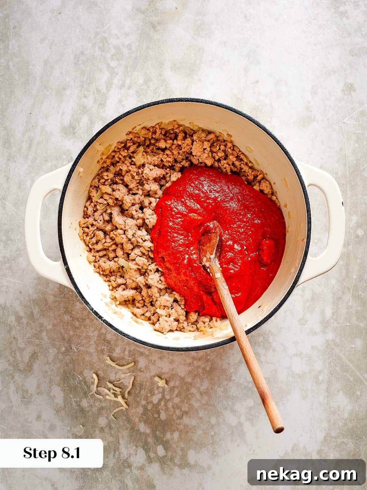 A generous spoonful of vibrant tomato sauce is being meticulously added to the cooked ground chicken in a skillet, ready to simmer and meld into a rich, savory base.