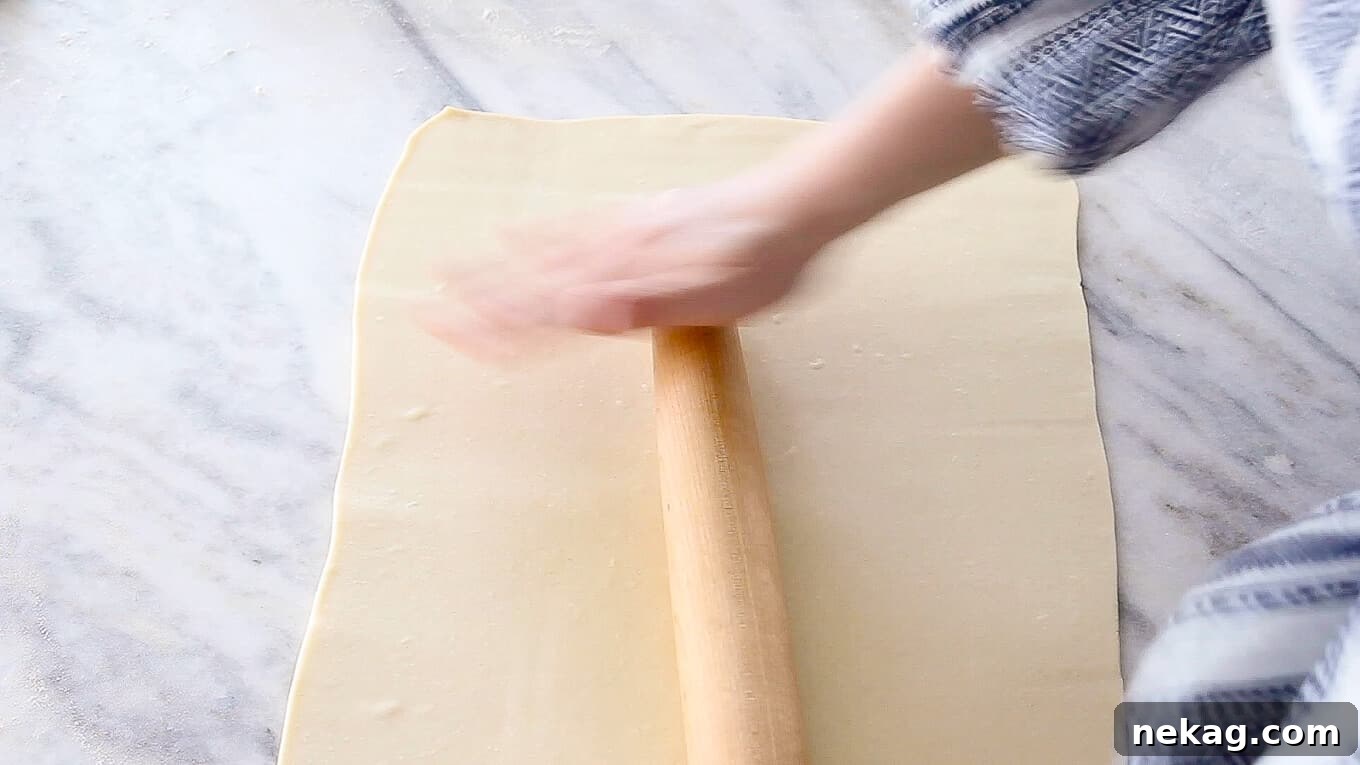 A baker expertly rolling out a sheet of inverse puff pastry dough with a wooden rolling pin on a clean surface