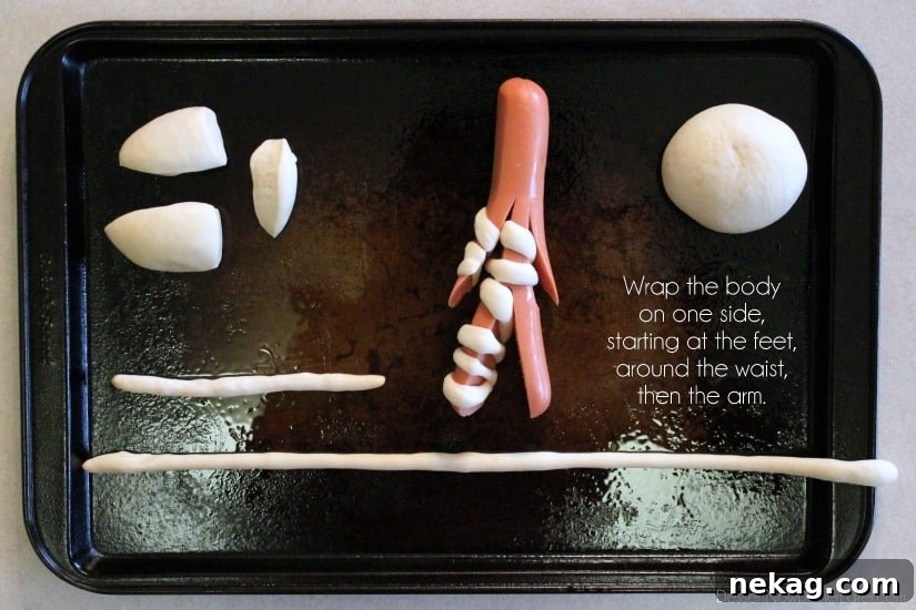 A hot dog mummy in progress, showing the first long dough strip meticulously wrapped around one leg, up the torso, and down one arm, illustrating the winding technique.