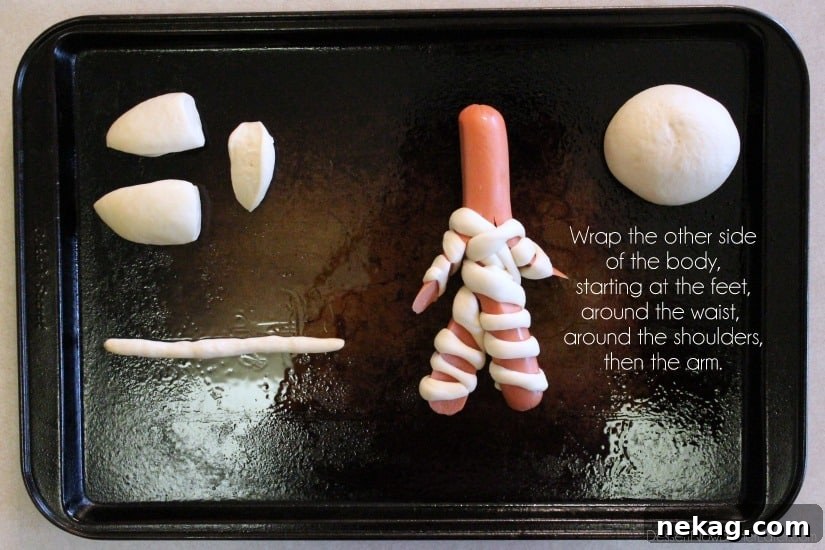 The hot dog mummy now fully wrapped on both sides, with dough strips covering both legs, the torso, and both arms, leaving only the head area exposed for the final wrapping step.