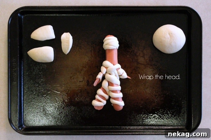A close-up of a hot dog mummy with its head expertly wrapped in a small dough strip, showcasing the small gap left for its eyes, ready for baking.