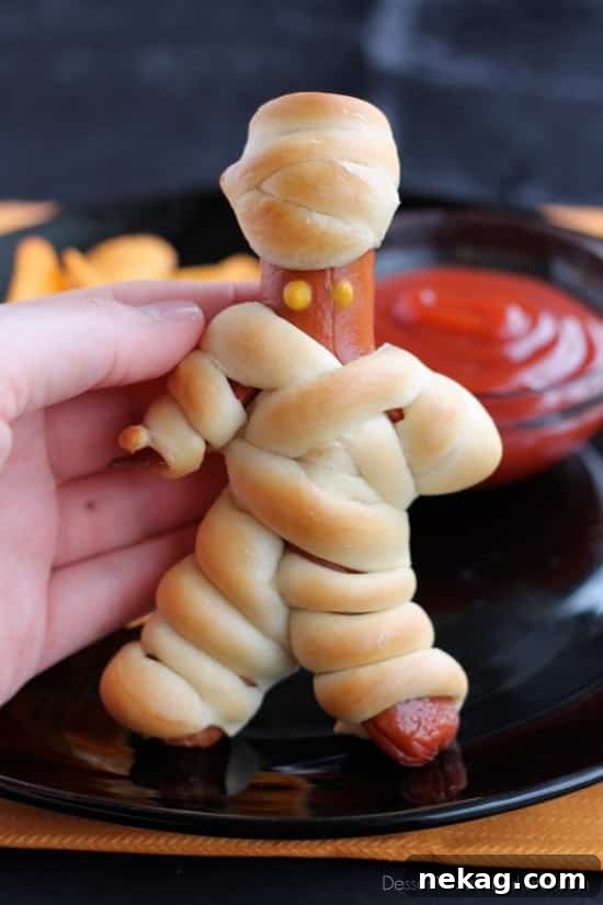 A full tray of freshly baked, golden brown Hot Dog Mummies, each with two mustard eyes, showcasing the finished product ready for a Halloween feast.