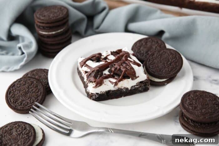 Fudgy Cookies and Cream Brownies 12 A close-up shot of a single Cookies and Cream Brownie on a plate, showcasing its soft, fudgy texture, studded with Oreo pieces and topped with creamy marshmallow frosting and a rich fudge drizzle.