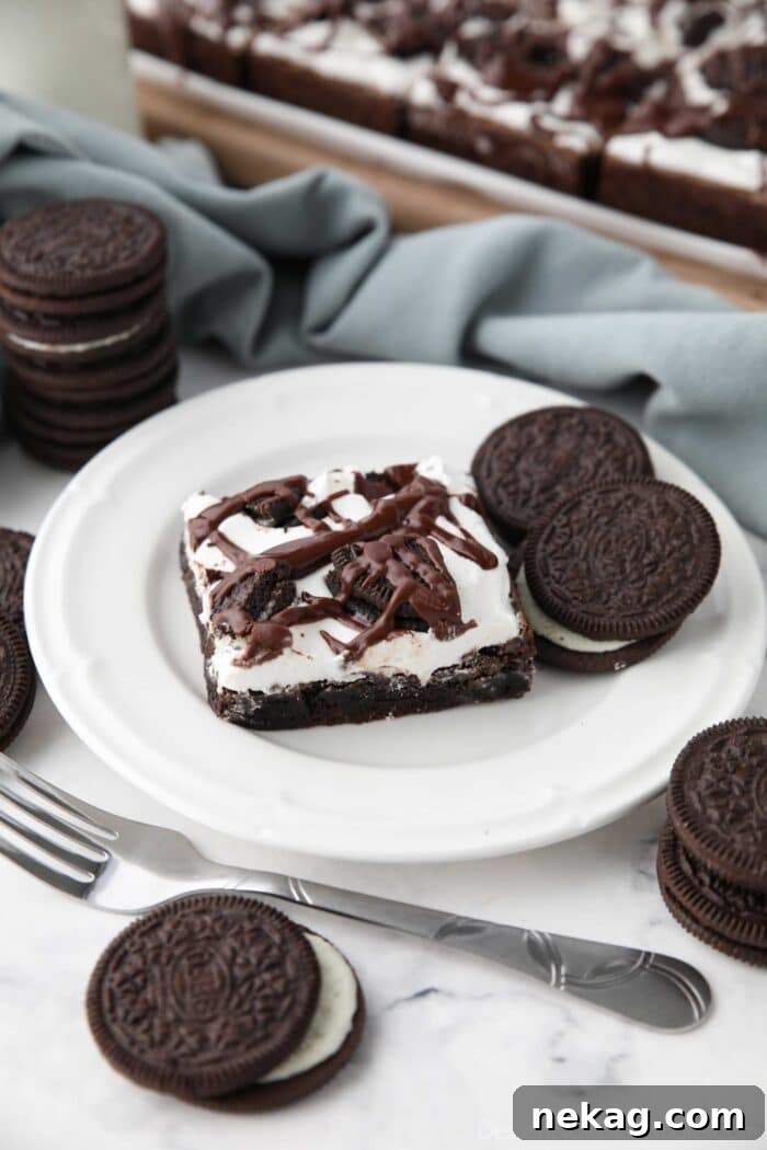Fudgy Cookies and Cream Brownies 3 A perfectly portioned Cookies and Cream Brownie rests on a plate, adorned with a swirl of marshmallow frosting, crushed Oreos, and a chocolate fudge drizzle, ready to be enjoyed.